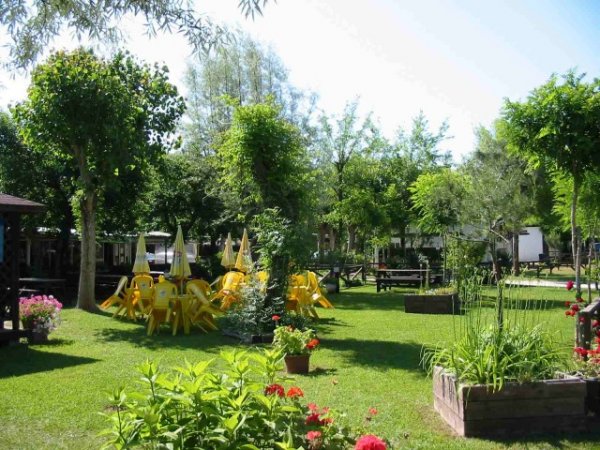 Giardino verde con sedie e tavoli gialli e ombrelloni, circondato da piante e alberi.