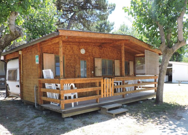 Casetta in legno con veranda, sedie bianche e circondata da alberi.