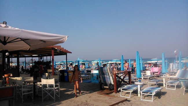 Una spiaggia affollata con ombrelloni blu e un chiosco con sedie da esterno.