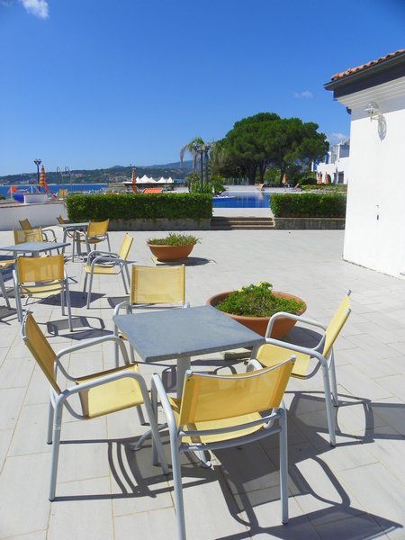 Tavoli e sedie gialle su un terrazzo, con piscina e alberi sullo sfondo.