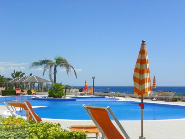 Piscina all'aperto con ombrellone arancione, vista mare e palme.