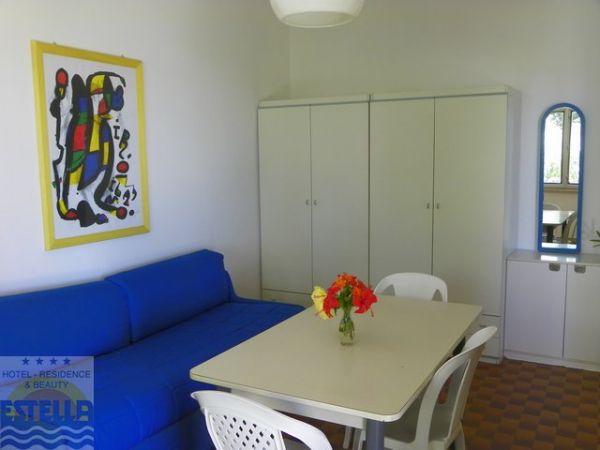 Interno semplice con divano blu, tavolo bianco, quattro sedie e un quadro colorato alle pareti.