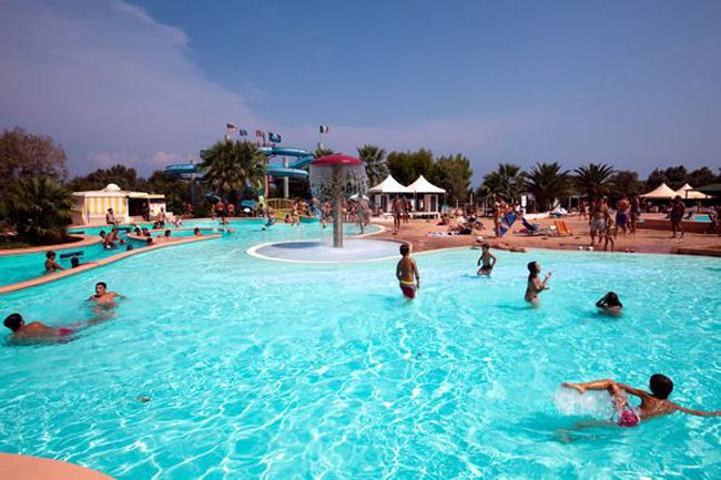 Piscina affollata con persone che nuotano e si rilassano, ombrelloni bianchi sullo sfondo.