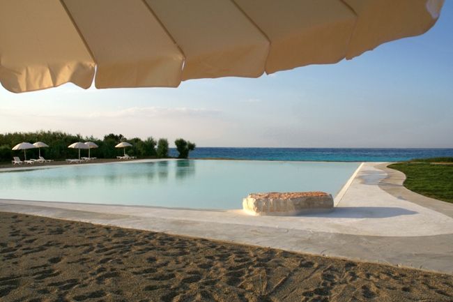 Piscina a sfioro vicino alla riva, con ombrellone beige e sabbia.