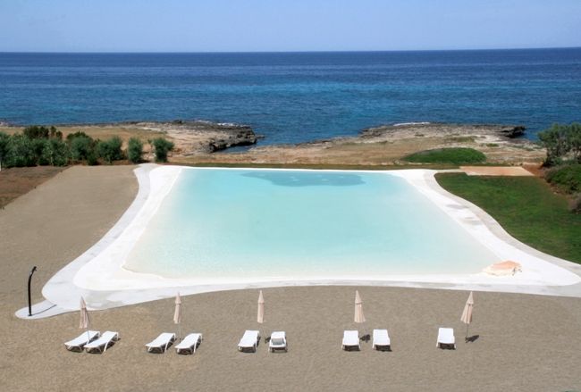 Una grande piscina vista dall'alto con lettini e ombrelloni bianchi e mare sullo sfondo.