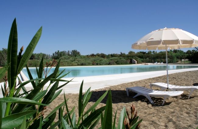 Lettini sotto un ombrellone beige accanto a una piscina, con piante in primo piano.