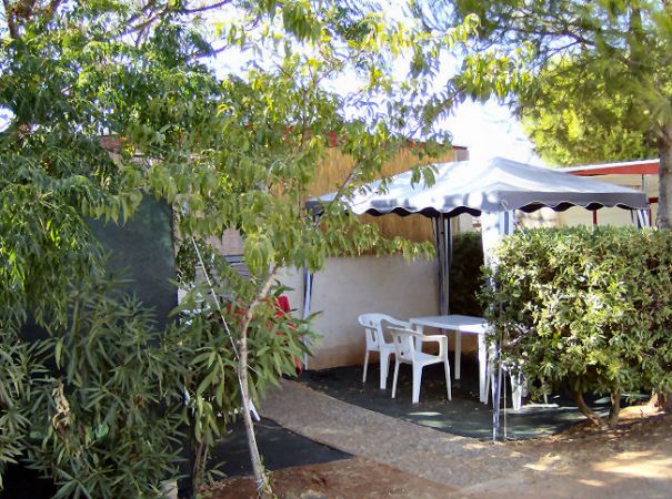 Una zona pranzo all'aperto con tavolo e sedie bianche sotto un gazebo tra alberi e arbusti.