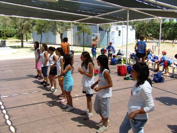 Persone in fila sotto una tettoia, su una superficie scoperta. Sembrano ascoltare istruzioni.