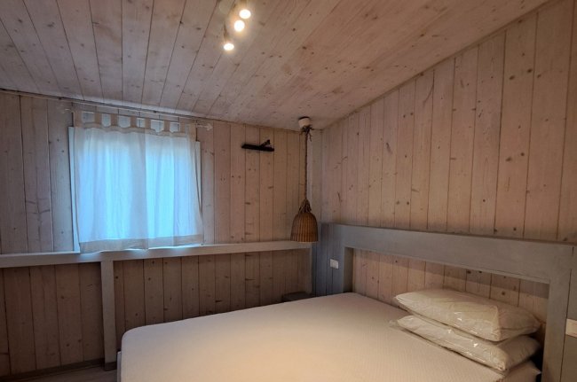 Camera da letto in legno chiaro con letto matrimoniale, finestra con tenda bianca e lampada appesa.