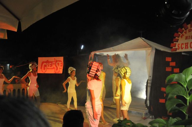 Ragazzi in scena con bigodini colorati, su un palco con il logo 