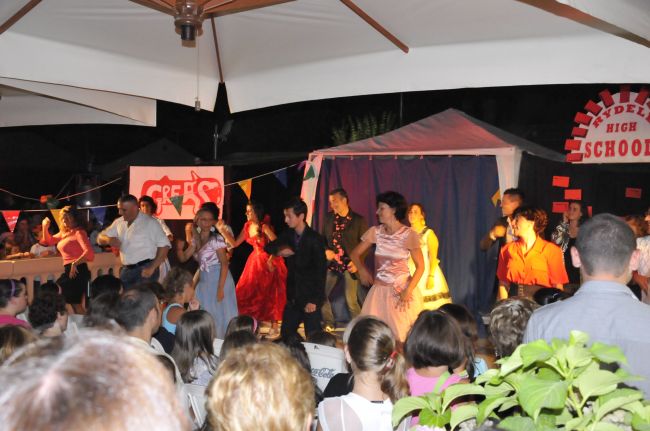 Persone in costume che ballano e cantano sul palco di un musical, con pubblico seduto.