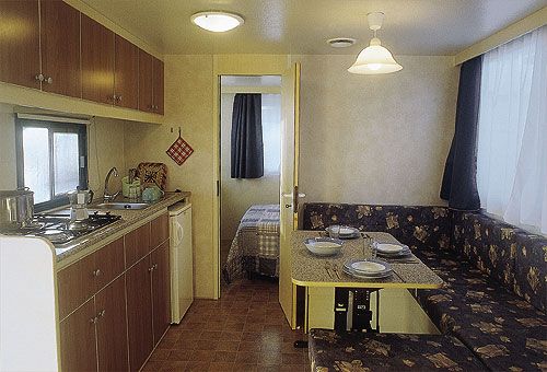 Interno di una roulotte con angolo cottura, sala da pranzo e letto in fondo.