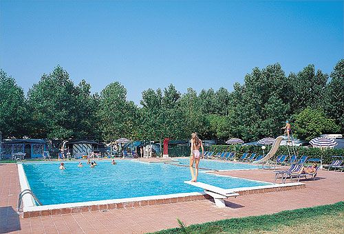 Una piscina all'aperto con diversi bagnanti, circondata da lettini e alberi.