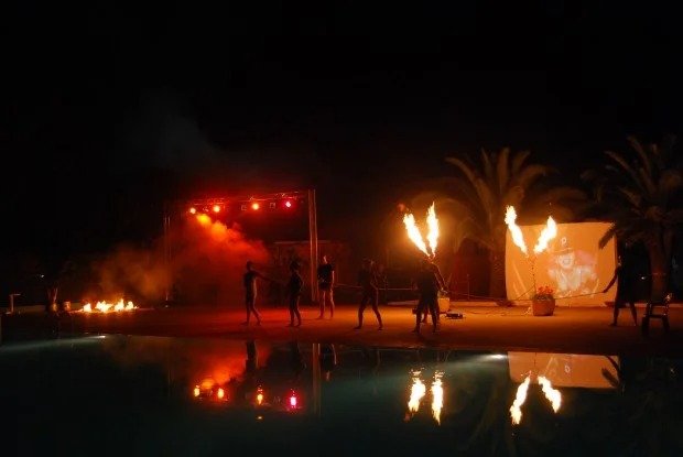 Spettacolo notturno con effetti di fuoco e luci rosse, palme sullo sfondo.