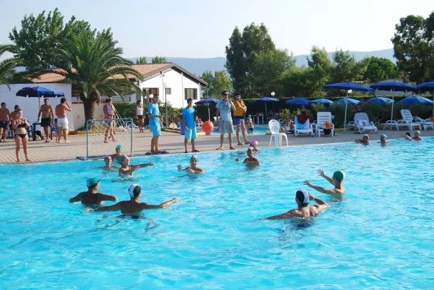Persone giocano in una piscina sotto un cielo sereno. Palme e sdraio sullo sfondo.