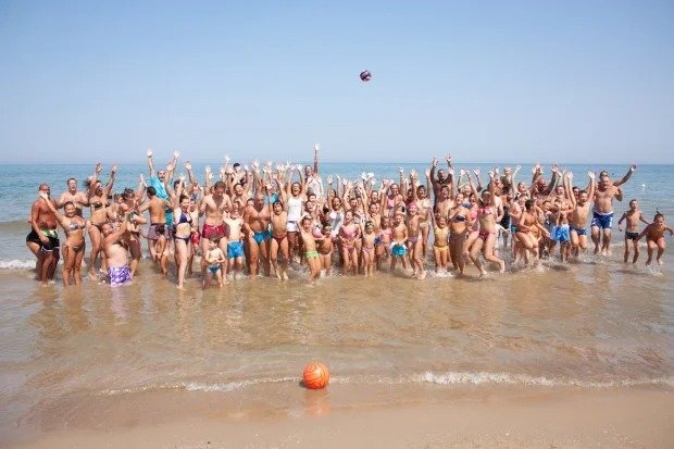 Un grande gruppo di persone salta gioiosamente in riva al mare con le mani alzate.