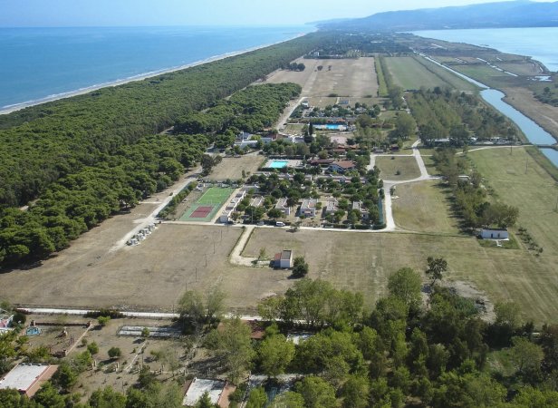 Veduta aerea di un villaggio turistico con piscine, campi sportivi e foresta lungo la costa.