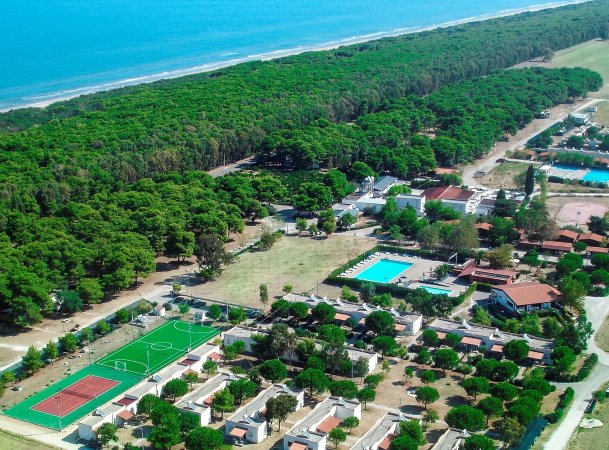 Vista aerea di un villaggio turistico con piscine, campi sportivi e una foresta vicino al mare.