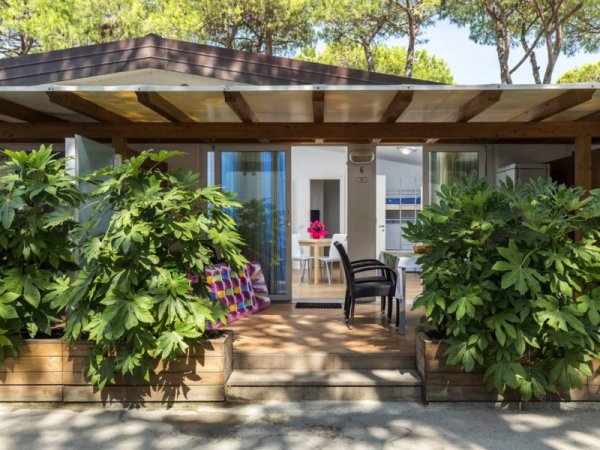 Bungalow con veranda coperta, piante verdi e tavolo interno visibile.