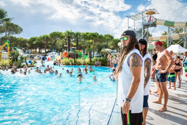 Persone in piscina seguite da figure in abiti sportivi e cappellini al bordo.