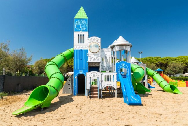Parco giochi a tema castello con scivoli verdi e blu su sabbia, cielo limpido e alberi sullo sfondo.