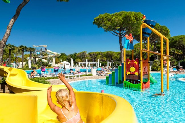 Scivolo giallo con una donna bionda, piscina con giochi d'acqua e resort in background.