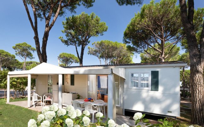 Casa mobile bianca con patio, sedie, e fiori bianchi. Alberi sullo sfondo.