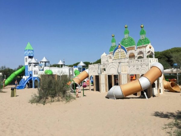 Parco giochi sulla sabbia con strutture a tema castello e scivoli colorati sotto un cielo azzurro.