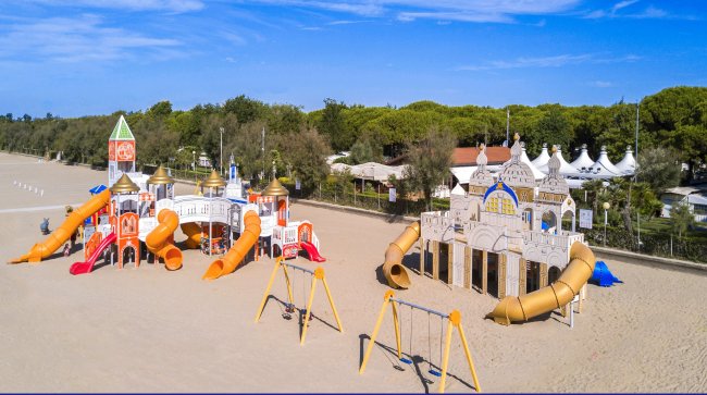Parco giochi sulla spiaggia con scivoli colorati e altalene, circondato da alberi e cielo blu.
