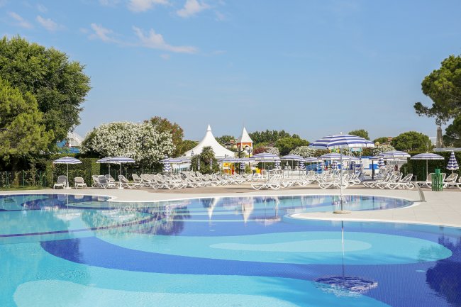 Piscina blu con lettini e ombrelloni bianchi e blu. Alberi e tende sullo sfondo.