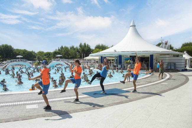 Animatori in uniforme arancione ed esercizi di ginnastica a bordo piscina affollata.